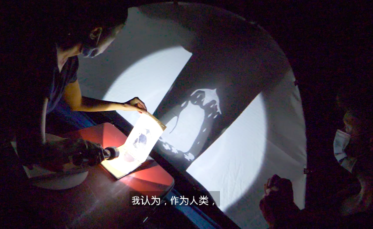 A flash light shines through paper projecting an image of melting woman on a white screen.
