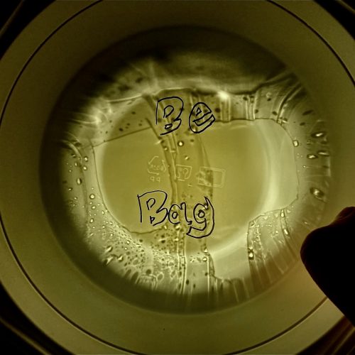 Upcycle 업사이클, Light shines through the bottom of a container illuminating beads of water. The word "UP" is cut out of the top side of the container. Recycle icons in the middle of the bottom part are seen through the top. The words "Be Bag" is written over in black marker or digital marker.