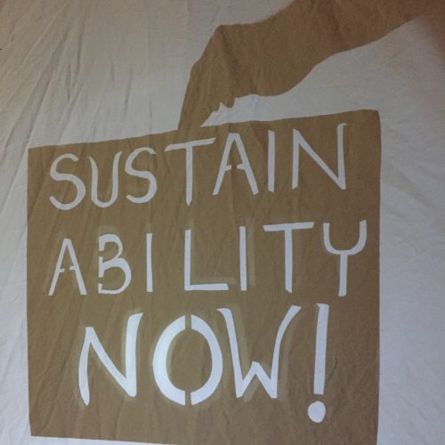 Sustainability, “Sustainability Now!” In white letters cut from a black shadow rectangle. Shadow of hand holds it from above.