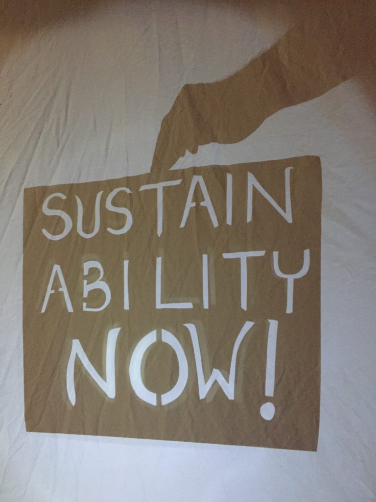 “Sustainability Now!” In white letters cut from a black shadow rectangle. Shadow of hand holds it from above.
