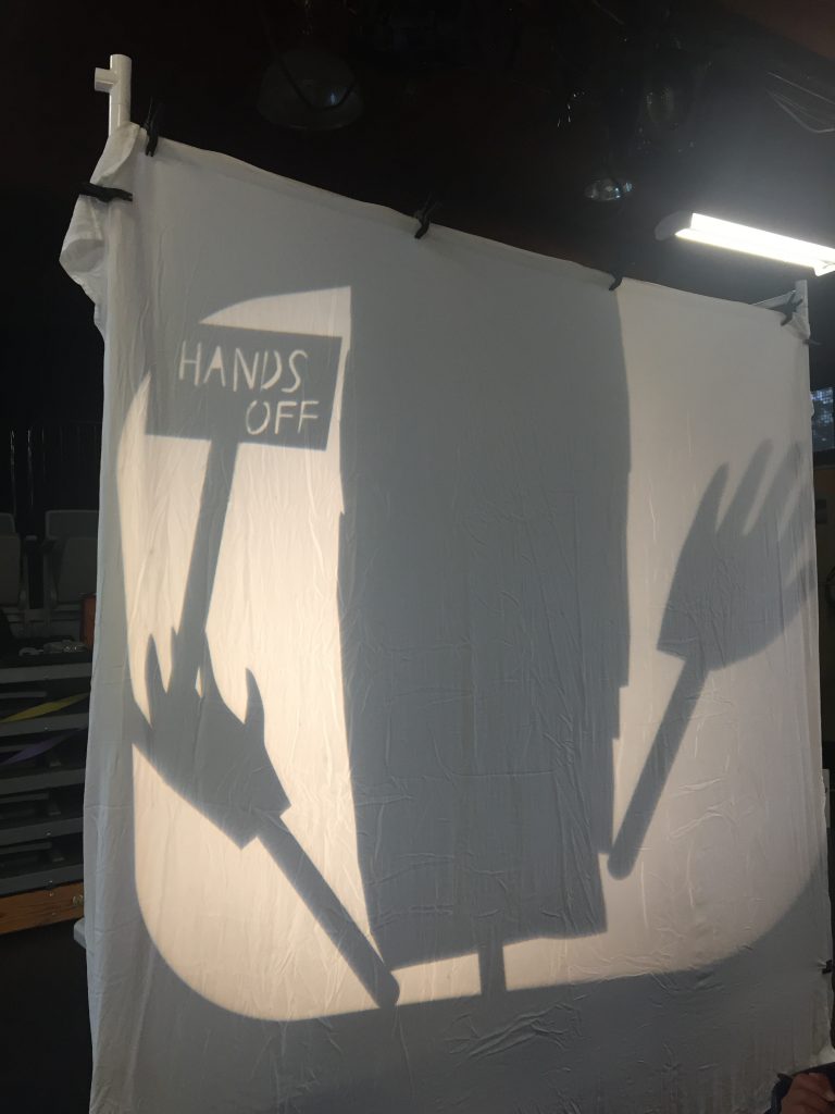 Two hands rise up on either side of a black vertical rectangle. The hand to the left holds a little sign the reads "Hands off."